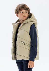 Beige gesteppte Weste mit Kapuze, mit steppdesign, einem Reißverschluss, seitlichen Taschen und getragen über einem marineblauen Langarmshirt.