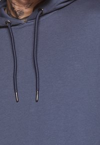 Närbild på en person som bär en blå huvtröja med snören och en nacktattoo av en ros.