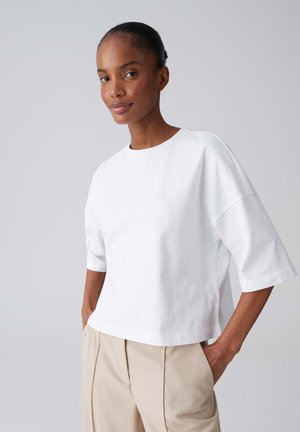 Vrouw met handen in de zakken, draag een losse witte korte mouw top en beige broek, staand tegen een effen lichtgrijze achtergrond.
