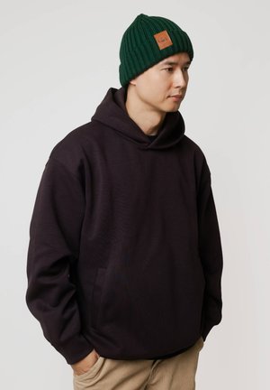 Ciemnofioletowa bluza z kapturem z przednią kieszenią oraz prążkowana zielona dzianinowa czapka z beżową skórzaną naszywką. Prosta, swobodna sylwetka.
