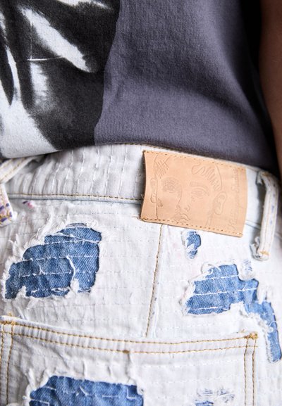 Distressed white denim shorts featuring blue patches, a textured leather label with a face illustration, and visible stitching details.