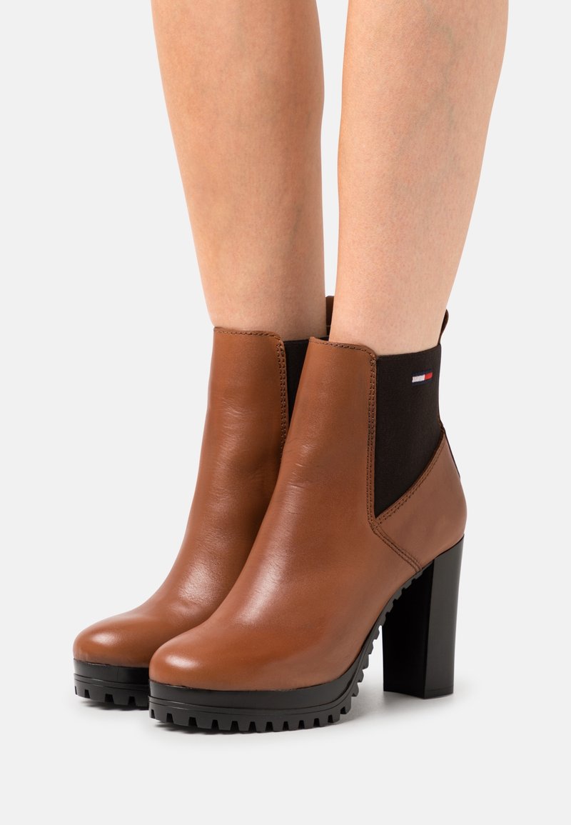 Bottines en cuir marron montant à la cheville avec des panneaux latéraux élastiques noirs, des semelles épaisses à crampons et des talons carrés, portées sur des jambes nues.