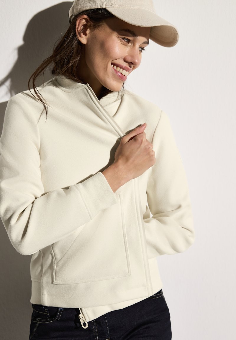 Veste en polaire blanche à zip avec un col montant, poches latérales et poignets côtelés, assortie d'une casquette beige clair sur fond neutre.