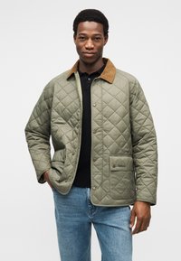 Πράσινο μπουφάν με καπιτονέ σχέδιο, καφέ γιακά από κορντόρι, κλείσιμο με κουμπιά τύπου snap και μπροστινές τσέπες. Φοριέται πάνω από μαύρο πόλο πουκάμισο και μπλε τζιν.