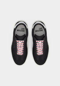 Ζευγάρι μαύρα sneakers Love Moschino με λευκές σόλες και λευκά κορδόνια, διακοσμημένα με κόκκινα σχέδια καρδιάς, σε θέα από πάνω.