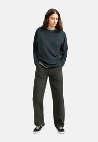 Felpa verde scuro con maniche raglan, indossata sopra una camicia verde chiaro, abbinata a pantaloni cargo larghi verde scuro, dotati di tasche.