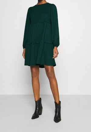 Kvinde iført en mørkegrøn, langærmet kjole med lag og indsnørrede skuldre samt sorte ankelstøvler, stående foran en ensfarvet baggrund.