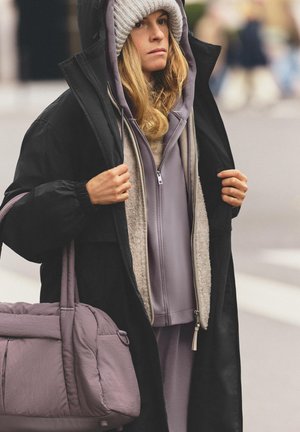 Long black coat over a grey zip-up hoodie, paired with light purple pants and a textured grey beanie. Carrying a soft purple bag.