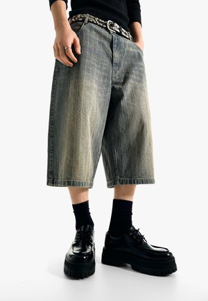 Person trägt weit geschnittene graue Jeansshorts, schwarzen Nieten-Gürtel, schwarze Socken und klobige schwarze Schnürschuhe vor weißem Hintergrund.