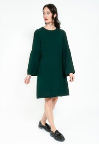 Zelena haljina do koljena s raširenim rukavima i okruglim izrezom. Izrađena od glatkog materijala, s opuštenim krojem. Uparena s crnim cipelama.