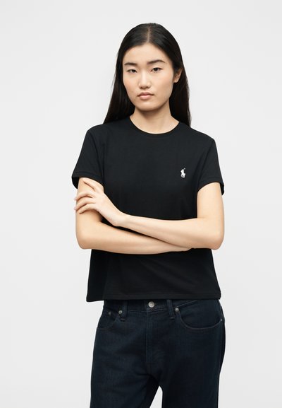 Νεαρή γυναίκα με μακριά μαύρα μαλλιά, ντυμένη με μαύρο κοντομάνικο μπλουζάκι με λογότυπο σε λευκό χρώμα και σκούρο τζιν, όρθια με τα χέρια σταυρωμένα.