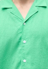 Κοντινό πλάνο ενός ατόμου που φοράει πουκάμισο ανοιχτού πράσινου χρώματος με γιακά και τρία λευκά κουμπιά ορατά.