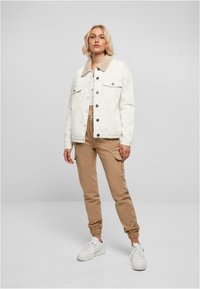 Veste en denim blanc avec un col en fourrure beige, deux poches poitrine, associée à un pantalon cargo beige et des baskets blanches. Design simple et couleurs neutres.
