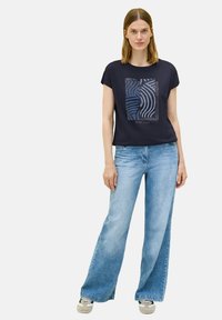 Camiseta de algodón color azul marino con un diseño gráfico abstracto en azul y negro, combinada con jeans acampanados de lavado claro y zapatillas con detalles en verde.