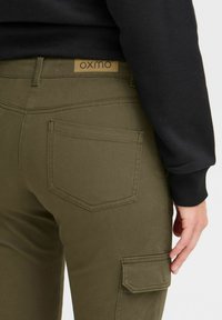 Pantaloni cargo verde oliva con una texture liscia, dotati di una tasca posteriore e un'etichetta in pelle marrone con la scritta 'OXMO' in vita.