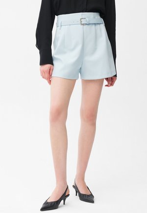 Vrouw draagt lichtblauwe high-waisted shorts met ceintuur, zwarte blouse met lange mouwen en zwarte slingback kitten heels, staand tegen een witte achtergrond.