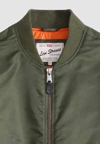 Olivengrønn bomberjakke med ribbet krage, glidelås foran og lys oransje innerfôr, Levi Strauss-merke synlig inne i kragen.