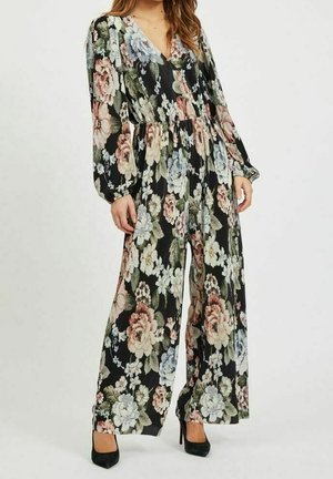 Nainen, joka pukeutunut pitkähihainen, leveälahkeinen kukkakuosinen haalari ja mustat korkokengät, seisoo yksinkertaisen vaalean taustan edessä.