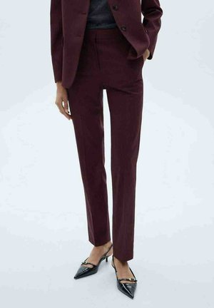 Person wears burgundy tailored pants and blazer with black pointed-toe slingback heels, standing with one hand in pocket.