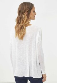 Top de punto blanco de mangas largas con un ajuste holgado y un diseño texturizado y transparente. Presenta un escote amplio y un dobladillo redondeado, combinado con jeans azules.