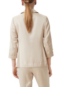 Beige tailored blazer with three-quarter sleeves, a notched collar, and rolled cuffs. Soft texture and clean lines enhance the minimalist design.