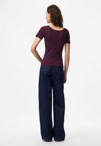 Moteris su garbanotais plaukais, vilkinti trumpomis rankovėmis purpurinį marškinius ir tamsiai mėlynas plačias kelnes su aukštu liemeniu, stovi atsukusi nugarą prieš baltą foną.