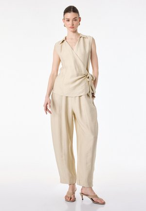 Donna che indossa un top beige senza maniche a portafoglio con cintura in vita, pantaloni ampi abbinati e sandali beige con tacco, in piedi su sfondo bianco.