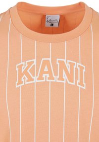 T-shirt de couleur pêche avec des rayures verticales blanches et le mot "KANI" en lettres grasses au centre. Matériau en coton doux et design à col rond.