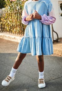 Robe en denim bleu avec des volants en étagère, manches longues en tie-dye roses et lavande. Baskets blanches avec des accents de boucle beige et chaussettes côtelées.