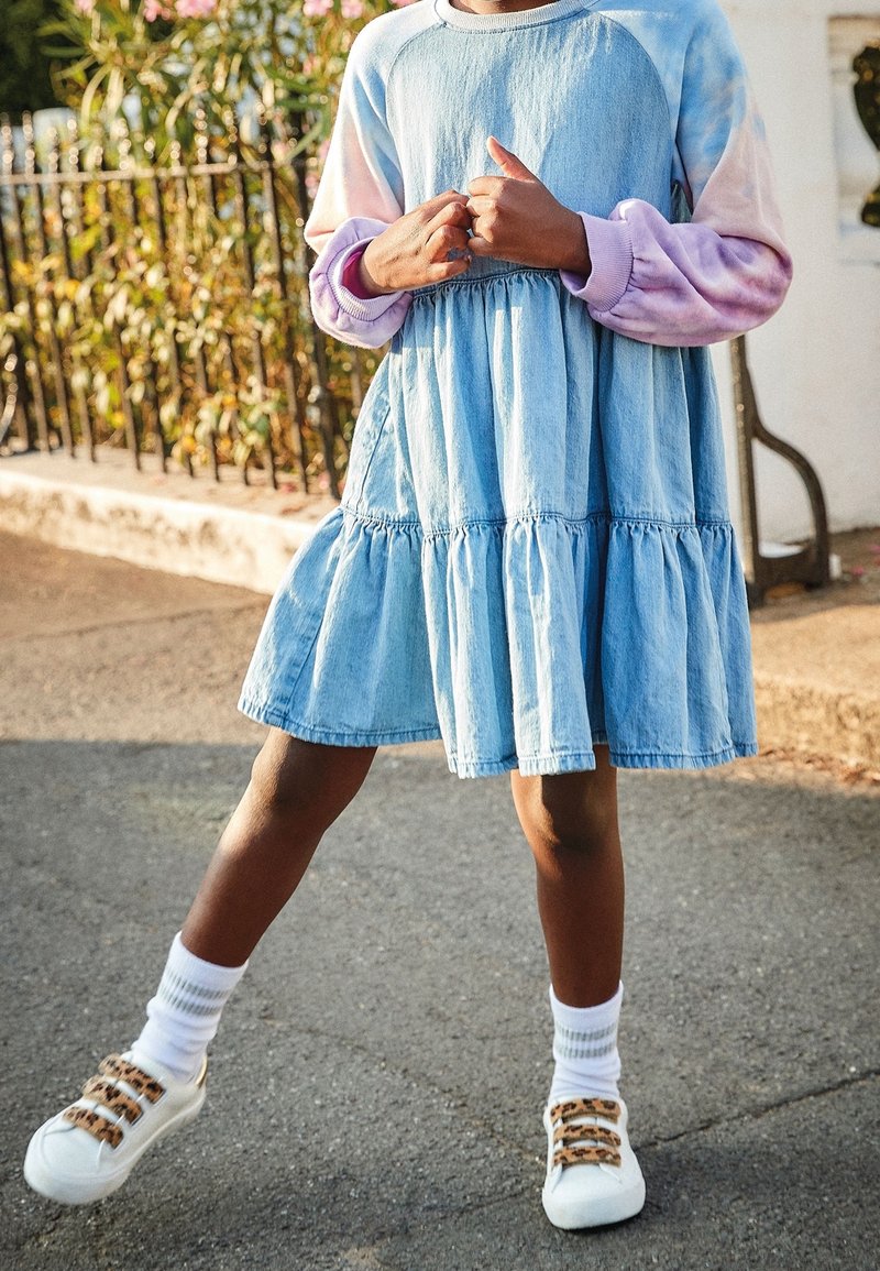 Robe en denim bleu avec des volants en étagère, manches longues en tie-dye roses et lavande. Baskets blanches avec des accents de boucle beige et chaussettes côtelées.
