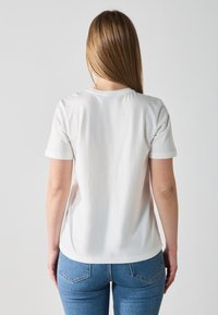 Vit bomulls T-shirt med korta ärmar och rundad halsringning, sedd från baksidan. Blå denimjeans med klassisk femfickorsdesign.