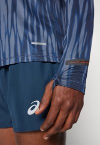 Camisola de manga longa Asics Actibreeze em azul com um padrão de ondas e buracos para os polegares; combinada com calções azul-marinho com um logótipo branco.