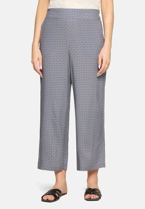 Vrouw met een high-waist, wijde broek met een blauw geometrisch patroon, gecombineerd met zwarte slip-on sandalen.