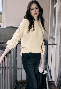 Krémový pletený svetr s texturovaným vzorem a ohrnutými rukávy, kombinovaný s černými džíny. Model drží černou bundu a světle zbarvenou látku.