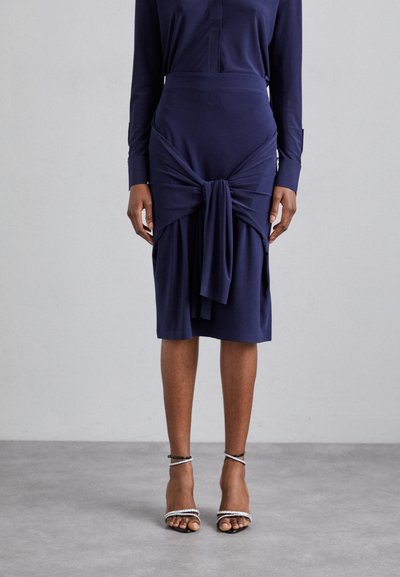 Marineblaues Midikleid mit gedrehtem Vorderteil, glatter Textur, langen Ärmeln und taillierter Silhouette, kombiniert mit metallic-farbenen, bandenartigen High Heels.