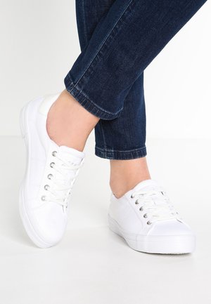 Persoon die witte lage vetersneakers en donkerblauwe spijkerbroek draagt, staand met gekruiste enkels op een witte vloer.