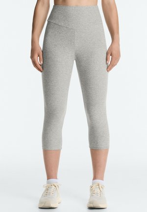 Femme portant des leggings gris clair courts et des baskets beiges sur un fond blanc uni.