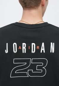 Μαύρο βαμβακερό μπλουζάκι με έντονα λευκά και πορτοκαλί γράμματα που γράφουν "JORDAN" και "23" στην πλάτη, με στρογγυλή λαιμόκοψη και υφή υφάσματος.