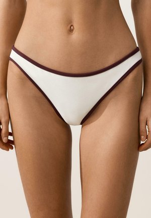 Trunchiul inferior al unei femei purtând chiloți de bikini albi cu margine maro închis, stând pe un fundal simplu.