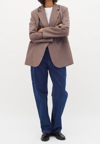 Persona in piedi con le braccia conserte, indossa un blazer marrone, una camicia bianca, jeans blu larghi e mocassini neri, su uno sfondo bianco semplice.