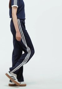 Marinträningsdräkt med vita ränder på sidorna. T-shirt med manschetter, byxor med avslappnad passform. Vita sneakers med bruna sulor.