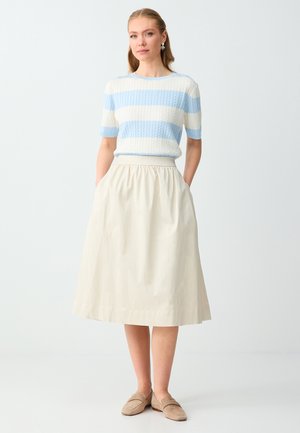 Vrouw met lichtbruin haar in een blauw-wit gestreept kortemouwtruitje, beige midi-rok en beige instapschoenen, staand met handen in de zakken.