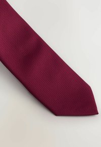 Burgundowa krawat o teksturowanym, plecionym wzorze. Wąski kształt z ostrym końcem. Gładka powierzchnia materiału, ułożona płasko na jasnym tle.