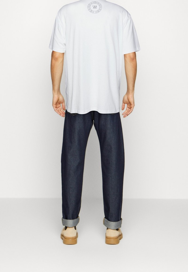 Bílý bavlněný tričko s kulatým výstřihem, v kombinaci s tmavě modrými džíny s volným střihem, s ohrnutými manžetami a béžovou obuví.