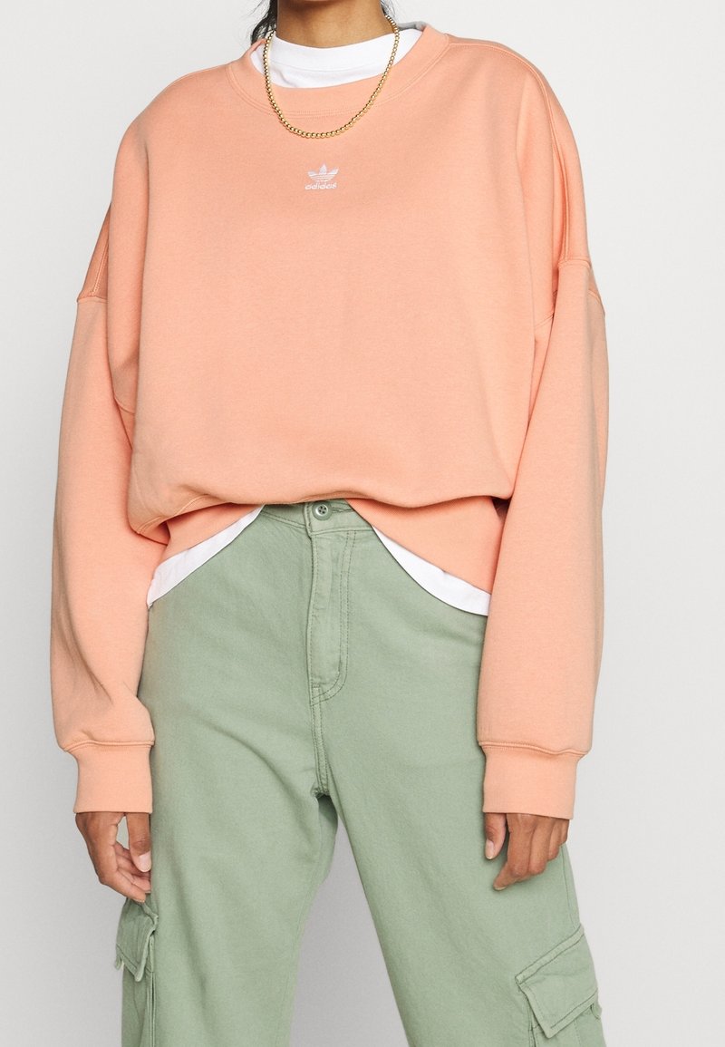 Person som bär en ljus persiko-färgad Adidas sweatshirt, vit undertröja, guldhalsband och ljusgröna cargobyxor med fickor.