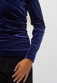 Top in velluto blu navy con maniche lunghe, caratterizzato da arricciature laterali e una silhouette aderente. Mano appoggiata su jeans neri.