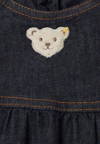 Denim mekko, jossa on kootut vyötärö, tummansininen kangas, oranssit ompeleet ja valkoinen brodeerattu karhukuosi rinnassa korvayksityiskohdalla.
