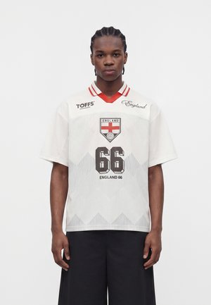 Mladý muž s vrkočmi nosí biely futbalový dres Anglicka s červeným golierom, emblémom Anglicka, číslom 66 a čiernymi šortkami, stojí nehybne.