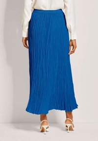 Frau in einer weißen Bluse, blauen plissierten Midirock und weißen High Heels, von hinten vor einer schlichten Wand gezeigt.