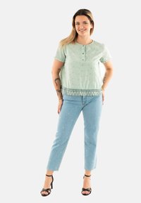 Blusa a maniche corte verde chiaro con dettagli ricamati e chiusura a bottone, abbinata a jeans blu chiaro con motivi sottili. Tacchi neri con cinturini.